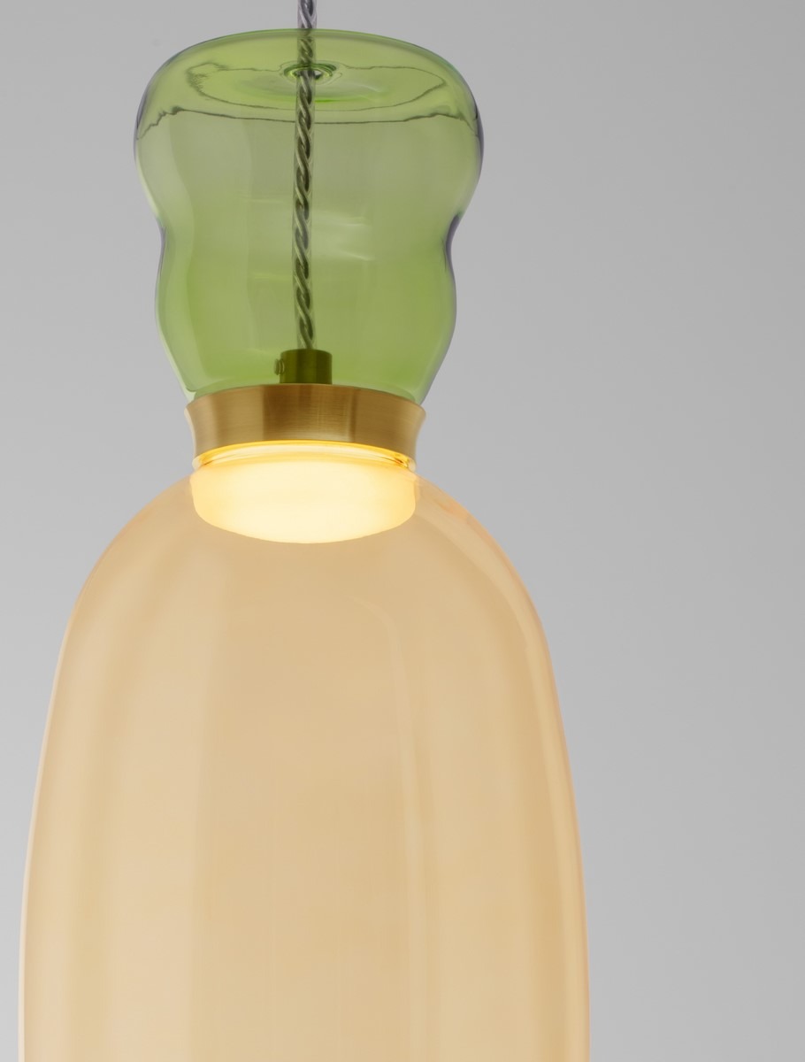 Lampa wisząca Perene z żółtą oprawą, posiadająca zielony klosz u góry. Zawiera jedno źródło światła LED 9W. Wisi na cienkim przewodzie, z metalowym wykończeniem u podstawy. Tło szare, podkreślające elegancki design lampy.