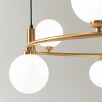 Lampa wisząca 6-punktowa w kolorze mosiężnym. Zawiera sześć okrągłych, matowych kloszy z białego szkła, zamocowanych na eleganckiej, złotej ramie. Ujęcie z boku, ukazujące szczegóły konstrukcji oraz kształt kloszy. Tło jasne, neutralne.