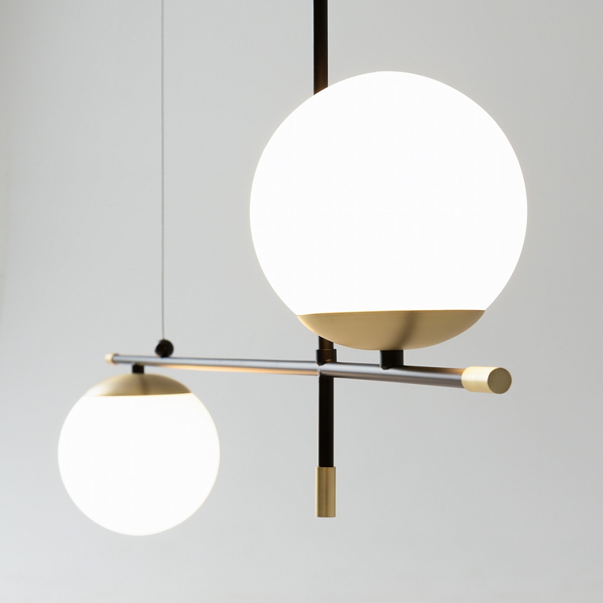 Lampa wisząca Nostalgia 2-punktowa w kolorze złotym. Dwie okrągłe, białe klosze zawieszone na czarnym stelażu, z metalowymi detalami. Klosze umieszczone na różnych wysokościach, tworzą nowoczesną kompozycję. Tło jasne, proste.