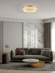 Lampa sufitowa Yotau 1xLED 70W w kolorze złotym, okrągła, z przezroczystym wzorem. Zainstalowana na suficie w nowoczesnym wnętrzu, otoczona szarymi zasłonami i eleganckim meblem, tworzy stylowy akcent oświetleniowy.