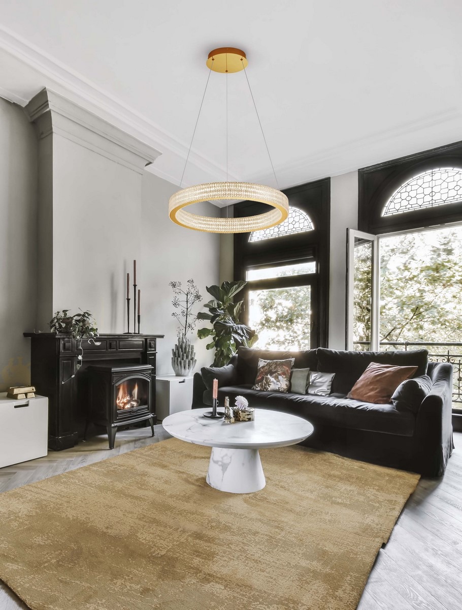 Lampa wisząca Yotau 1xLED 70W w kolorze złotym, okrągła, zawieszona na cienkich linkach. W tle elegancki salon z czarną kanapą, okrągłym stołem z marmurowym blatem oraz kominkiem. Przez okna wpada naturalne światło.