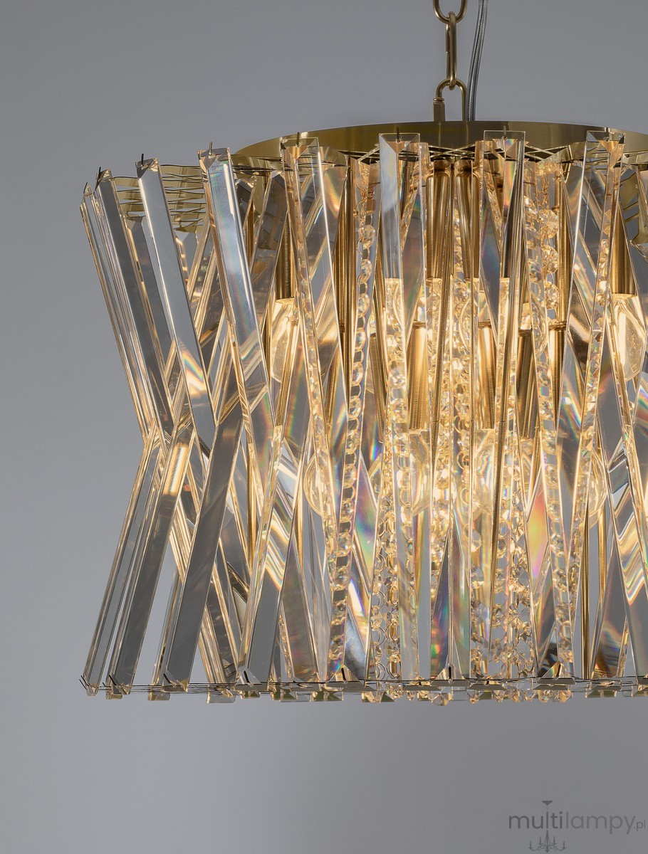 Lampa wisząca Melunto 8xE14 w satynowym mosiądzu. Ozdobna konstrukcja z przezroczystych, błyszczących elementów, układających się w geometryczny wzór. Zbliżenie na detale, widoczne żarówki w środku, wisząca na łańcuszku. Tło szare.