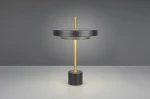 Lampa stołowa Keaton z czarnym kloszem i złotym stelażem, z okrągłą podstawą. Ujęcie frontalne, produkt w pozycji stojącej, emitujący światło. Wysokość stelaża podkreśla nowoczesny design. Tło szare, minimalistyczne.