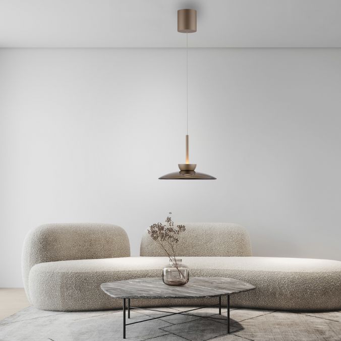 Brązowa lampa wisząca Pure Moon 2xLED 16W, z minimalistycznym kloszem, zawieszona na cienkim przewodzie. W tle widoczna nowoczesna kanapa i okrągły stół z dekoracyjną rośliną w szklanym wazonie, na jasnym tle.