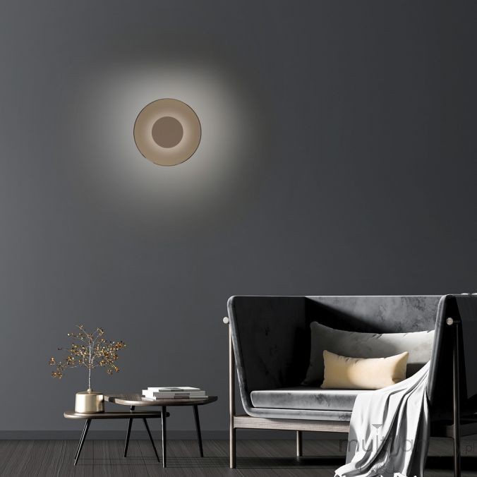 Kinkiet Pure Moon 2xLED 14W w kolorze brązowym, zamontowany na szarej ścianie. Okrągły kształt z centralnym otworem, emitujący delikatne światło, w tle widoczna szara sofa z poduszkami i stolik kawowy.