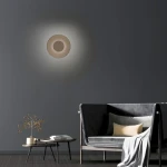 Kinkiet Pure Moon 2xLED 14W w kolorze brązowym, zamontowany na szarej ścianie. Okrągły kształt z centralnym otworem, emitujący delikatne światło, w tle widoczna szara sofa z poduszkami i stolik kawowy.