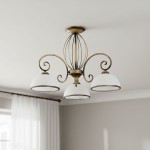 Żyrandol Indra 3-punktowy O2493 W3 PAT w kolorze złotym z trzema białymi kloszami. Produkt zamontowany na suficie, z eleganckimi, zakrzywionymi ramionami. Tło jasne, z miękkimi zasłonami w oknie.