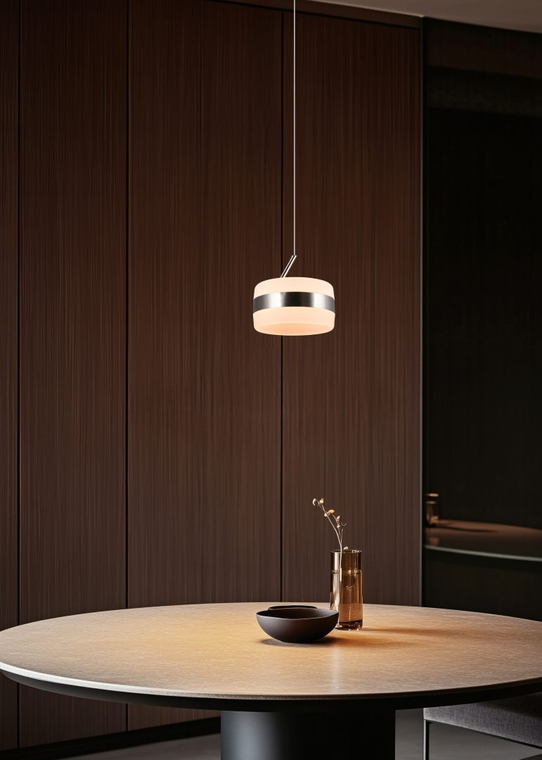 Lampa wisząca Dios z 1xLED 8W w kolorze niklowym, o okrągłym kształcie z matowym kloszem i metalowym wykończeniem. Zawieszona nad okrągłym stołem, w tle ciemne drewniane szafki. Długość zawieszenia podkreśla nowoczesny design.