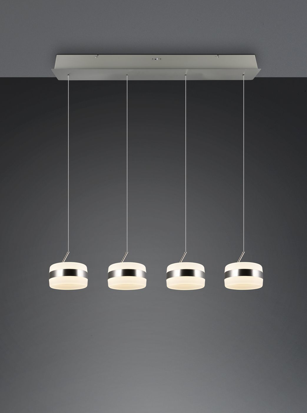 Lampa wisząca Dios z czterema źródłami LED, o mocy 32W, w kolorze niklowym. Okrągłe klosze z przezroczystym wykończeniem wiszą na cienkich linkach z metalowym stelażem, zawieszone na prostokątnej podstawie. Tło ciemnoszare.