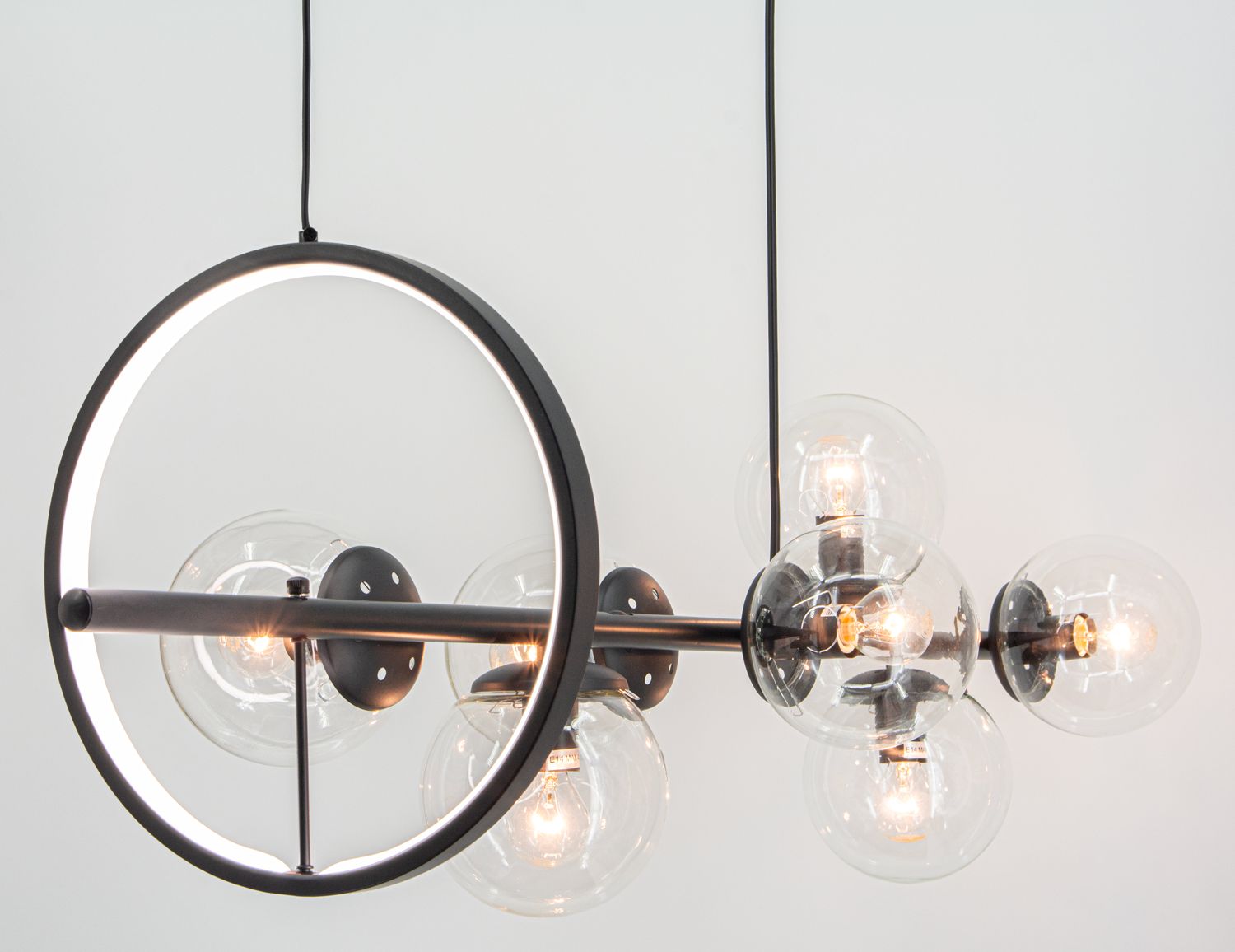 Lampa wisząca Galaxy w kolorze czarnym, z siedmioma gniazdami E14 oraz jednym LED. Okrągły, matowy metalowy element z białym podświetleniem i sześcioma przezroczystymi kloszami, w których znajdują się żarówki, zawieszona na długim kabelku.