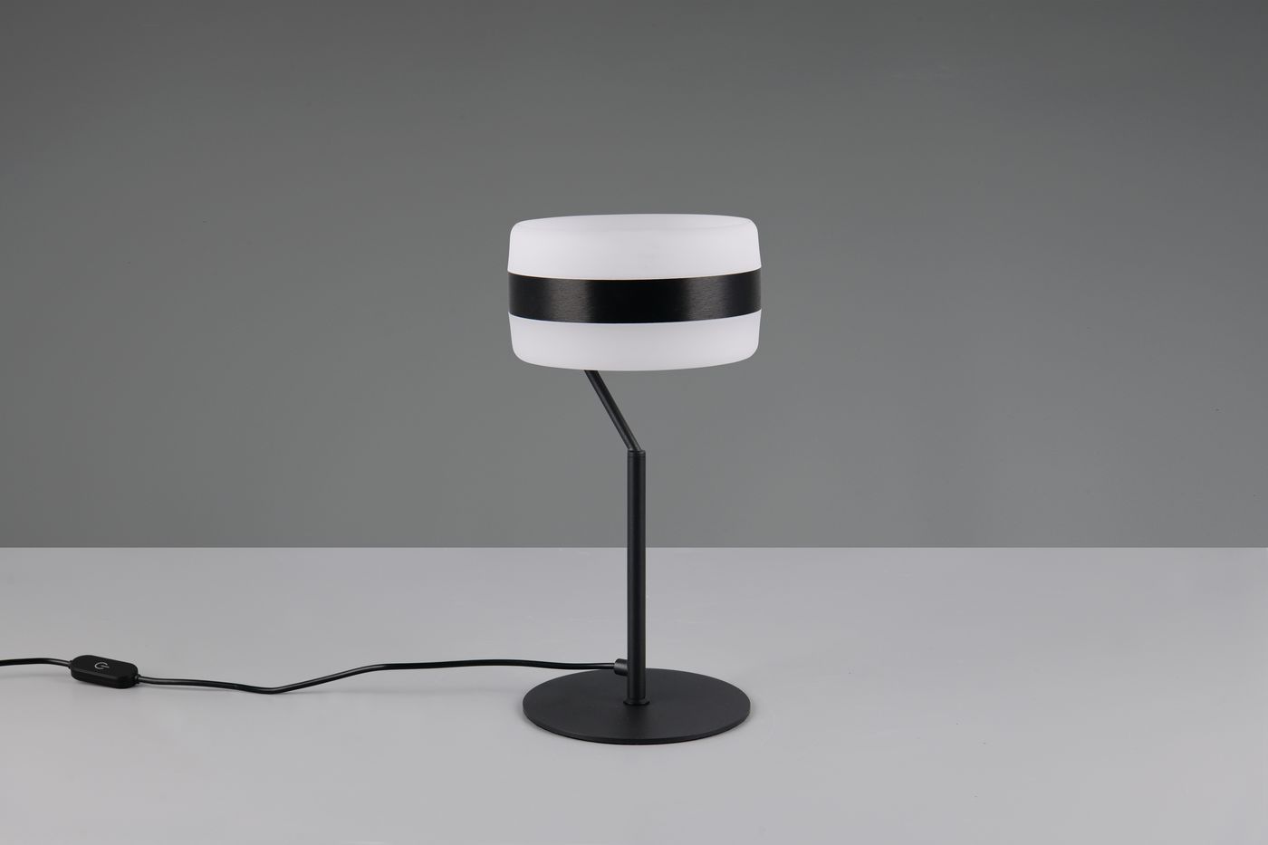 Lampa stołowa Dios z białym kloszem i czarnym paskiem, zamocowana na cienkiej czarnej nodze i okrągłej podstawie. Zasilana jedną diodą LED 8W, z kablem z włącznikiem na powierzchni. Ujęcie frontalne na jednolitym tle.