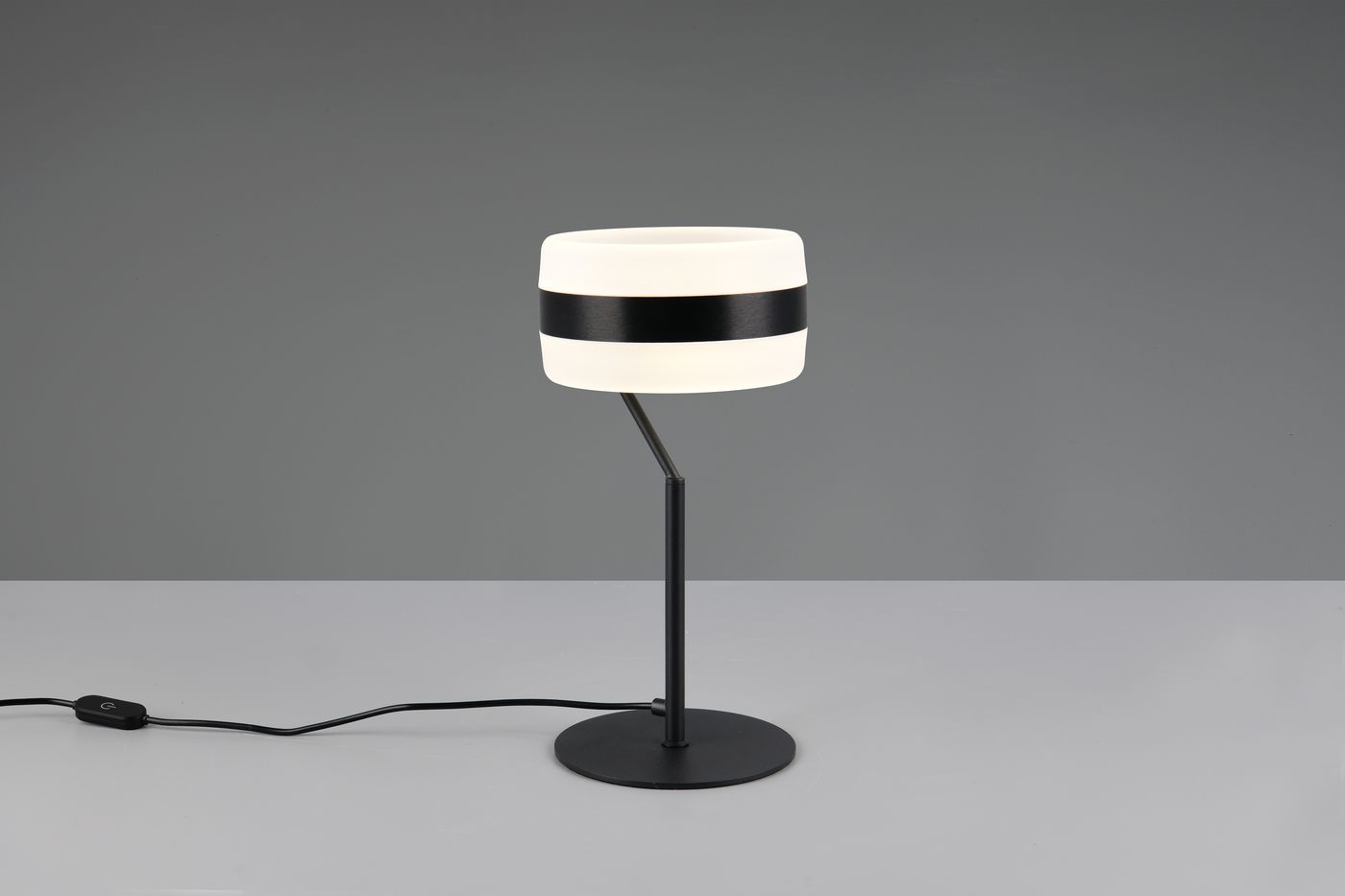 Lampa stołowa Dios z czarnym metalowym stelażem i okrągłą podstawą. Klosz w odcieniu białym z czarną poziomą linią. Produkt z kablem z przełącznikiem, ustawiony na stole, z widokiem z boku. Idealna do nowoczesnych wnętrz.