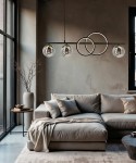Lampa wisząca Malmo z trzema szklanymi kloszami w czarnej i złotej kolorystyce, zawieszona nad szarym narożnikiem. W tle widoczna ściana w odcieniu brązu oraz duże okna. Elementy okrągłe w konstrukcji lampy.