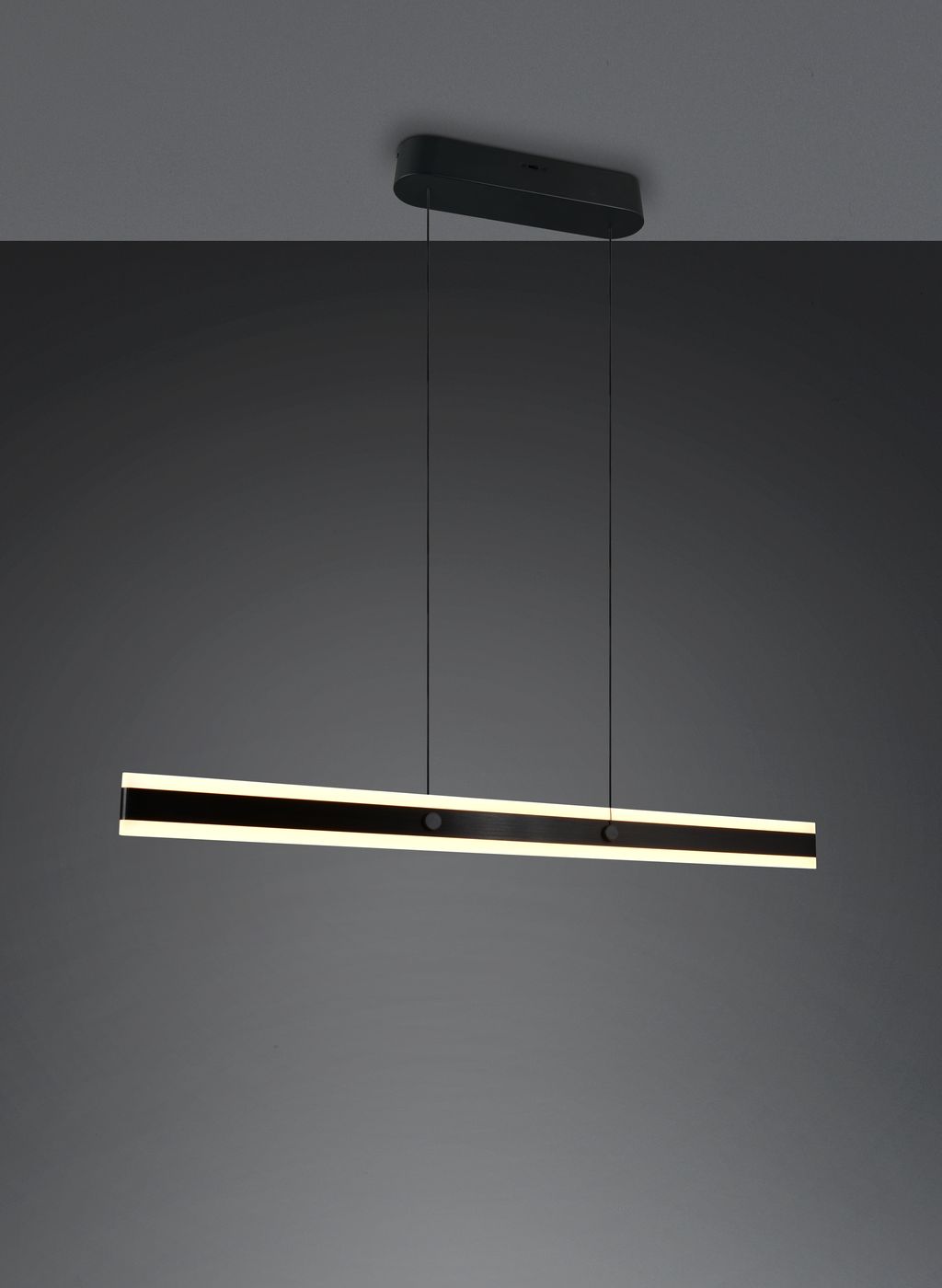 Lampa wisząca Arcola w kolorze czarnym, z wbudowanym źródłem LED 35W CCT. Prosty, długi kształt z białym podświetleniem na krawędziach, zawieszona na dwóch cienkich linkach. Montaż sufitowy na prostokątnej podstawie.