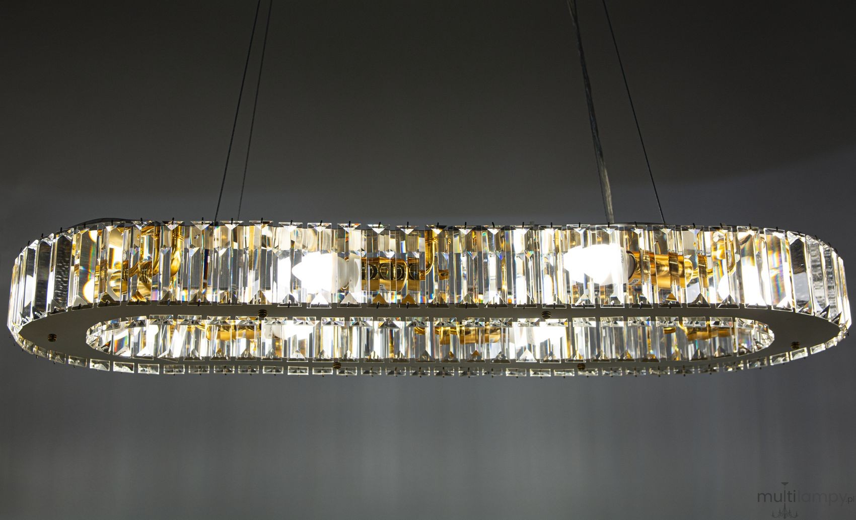 Lampa wisząca Windsor 8xE14 w kolorze złotym, o owalnym kształcie, z przezroczystymi kryształkami. Widoczna struktura z metalowymi elementami, zawieszona na cienkich linkach. Oświetlenie w środku zapewnia intensywne światło.