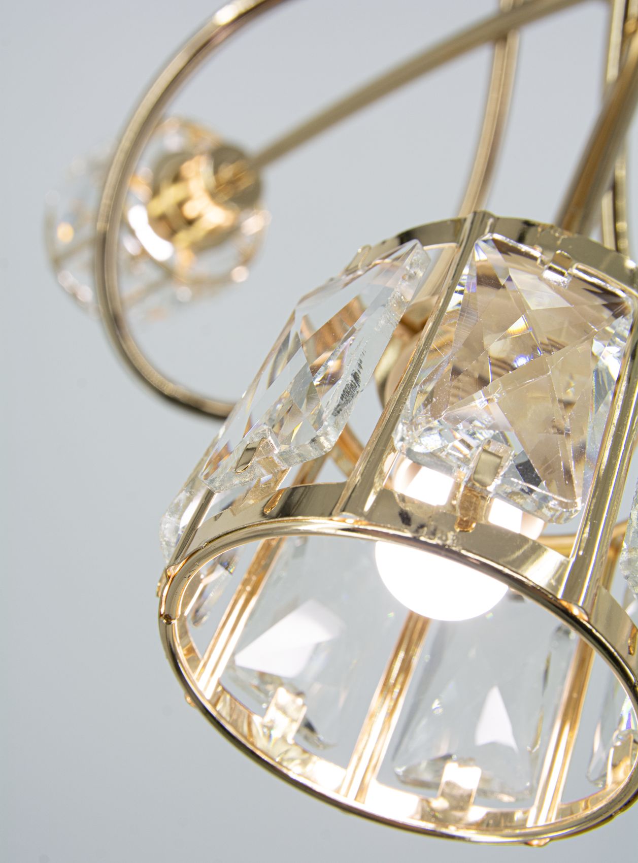 Lampa wisząca Aurora w kolorze złotym z trzema oprawkami E14. Elementy dekoracyjne z przezroczystego kryształu, o cylindrycznym kształcie, z metalową ramą. Widok zbliżeniowy na detale klosza oraz źródło światła.