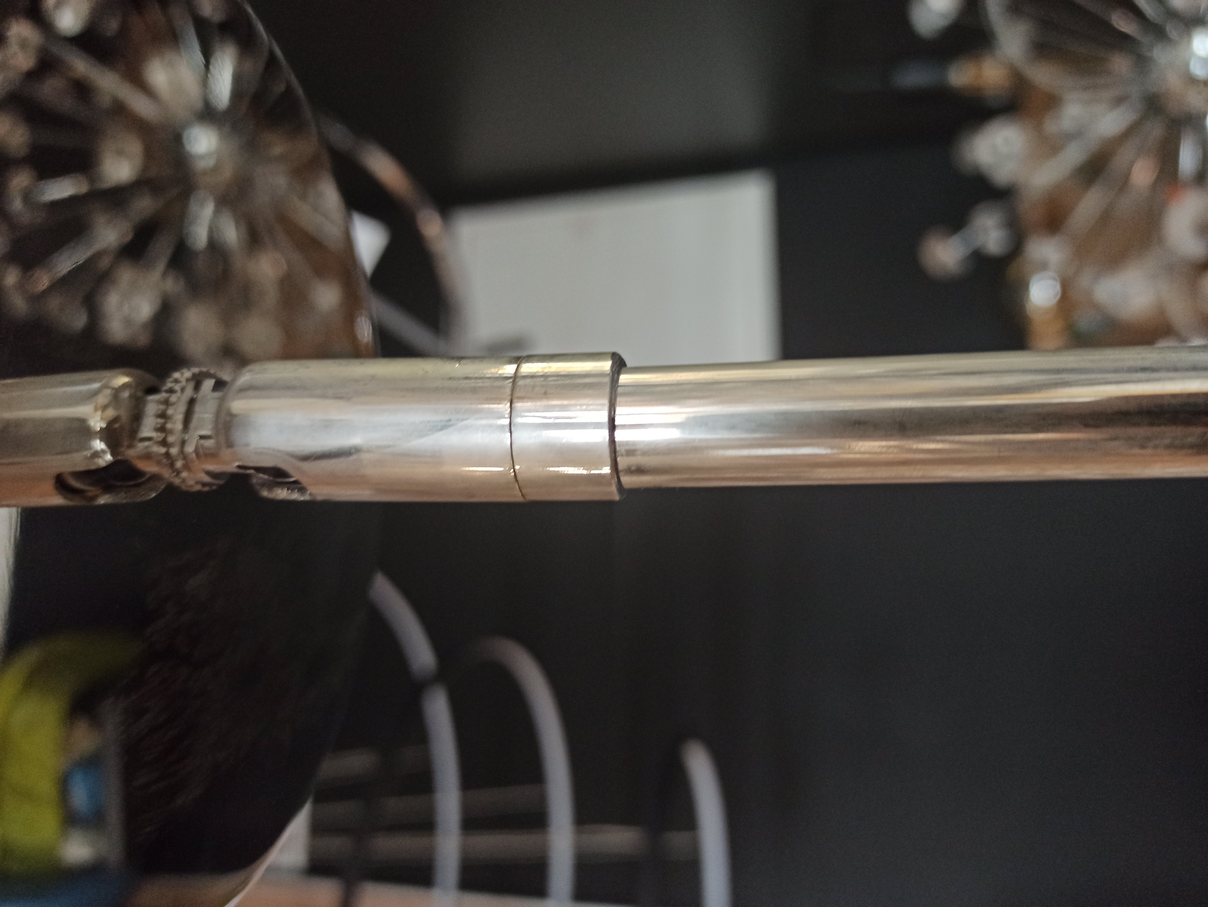 Lampa wisząca Oxford 4-punktowa, metalowa, z połyskującym wykończeniem. Ujęcie z bliska, pokazujące detale mocowania i rury, z tłem w odcieniach czerni. Elementy dekoracyjne lampa w tle są rozmyte.