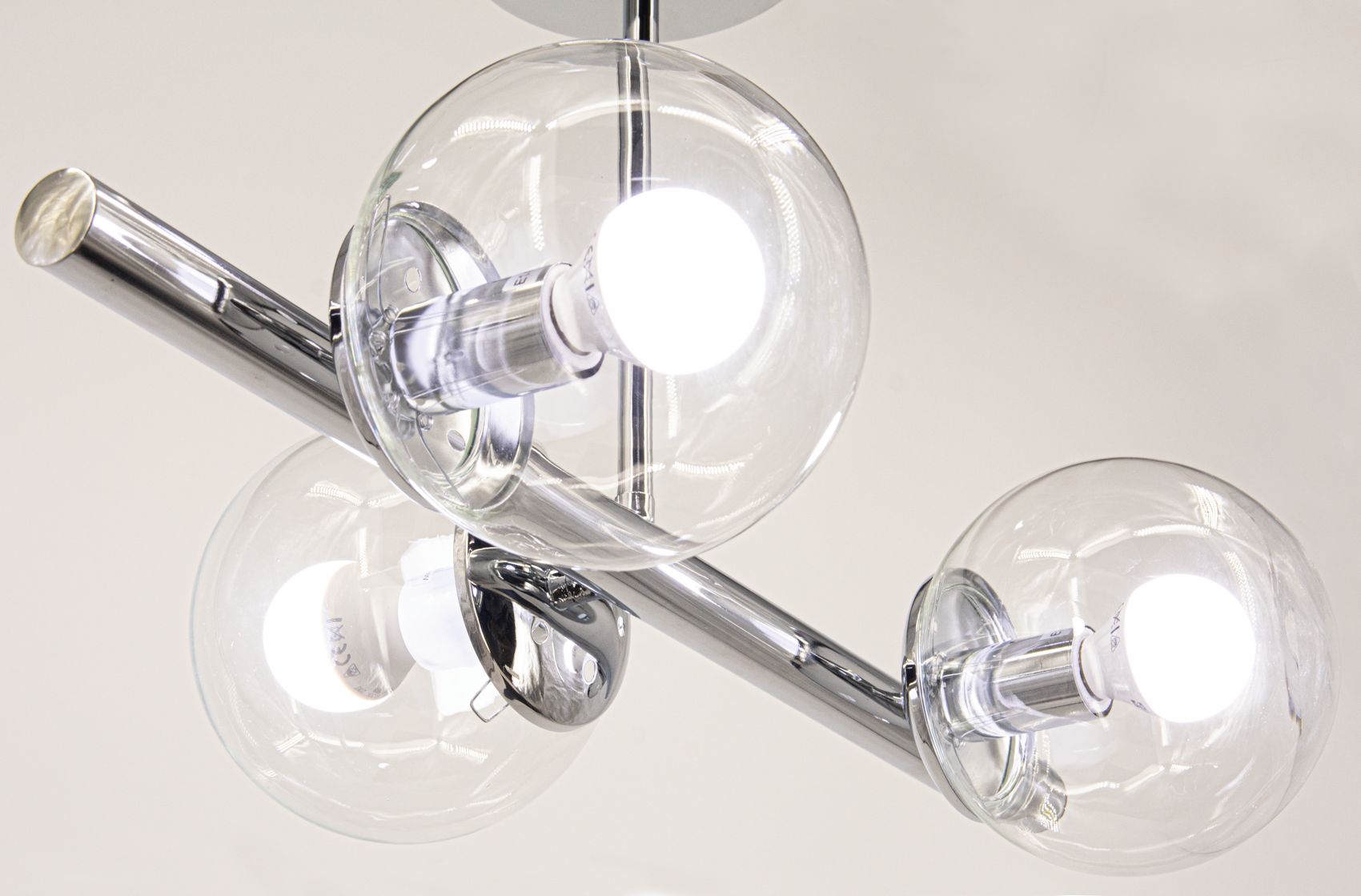 Lampa wisząca Otis z trzema kloszami w kształcie kul, wykonana z przezroczystego szkła. Chromowany metalowy stelaż, z widocznymi gniazdami E27. Kąt ujęcia z dołu, ukazujący detale konstrukcji i źródła światła. Tło jasne, minimalistyczne.