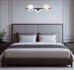 Lampa wisząca Otis 3xE27 w chromowanej obudowie, umieszczona nad łóżkiem w sypialni. Posiada trzy szklane klosze w kształcie kul, oświetlające przestrzeń. Tło stanowi jasna ściana z delikatnymi detalami, a meble są w odcieniach brązu.