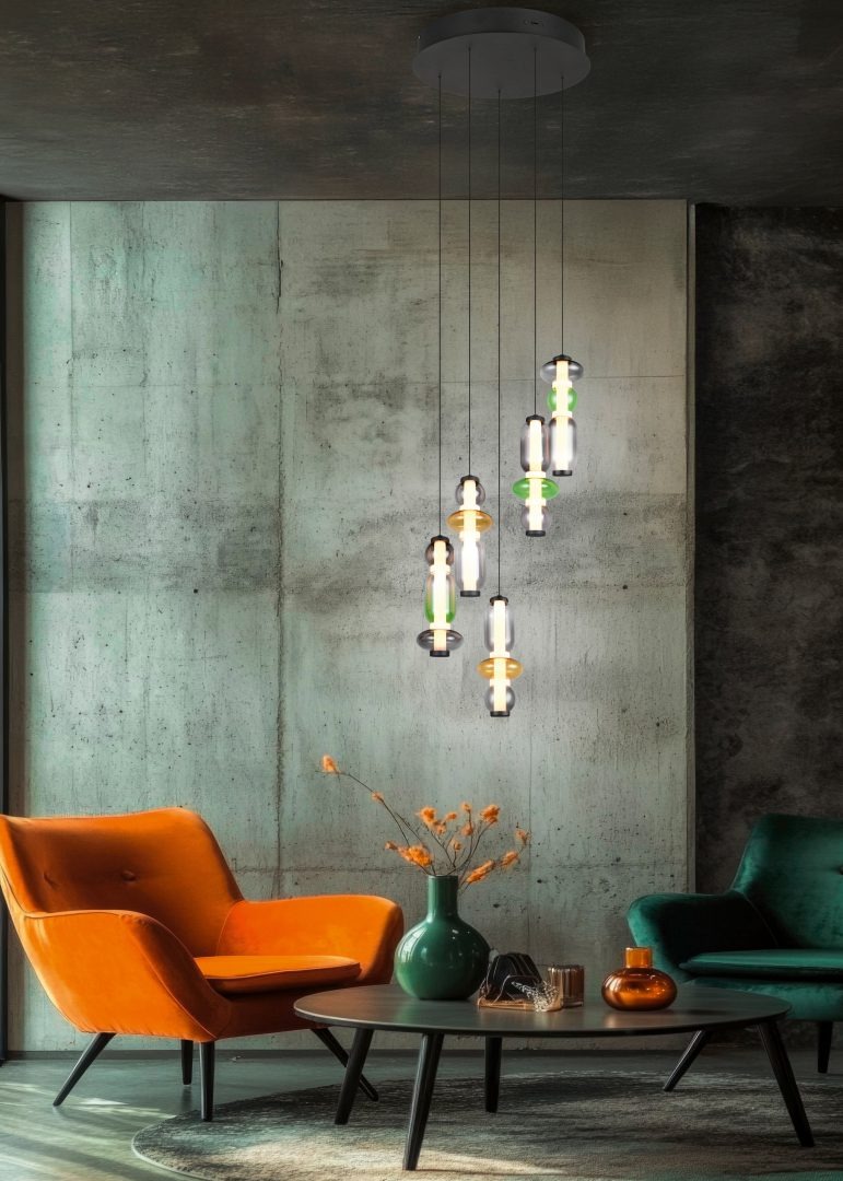 Lampa wisząca Minerva, czarna, z pięcioma źródłami LED 40W CCT, w multikolorowym wzornictwie. Zawieszona na cienkich przewodach, w nowoczesnym wnętrzu z pomarańczowym fotelem i zielonym wazonem. Tło w odcieniach szarości.