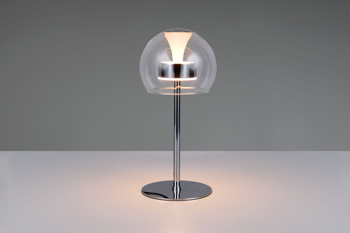 Lampa stołowa Flynt z jedną żarówką LED 8W, chromowana podstawa, przezroczysta klosz w kształcie kuli. Ujęcie frontalne, stojąca na białym blacie, emitująca ciepłe światło.
