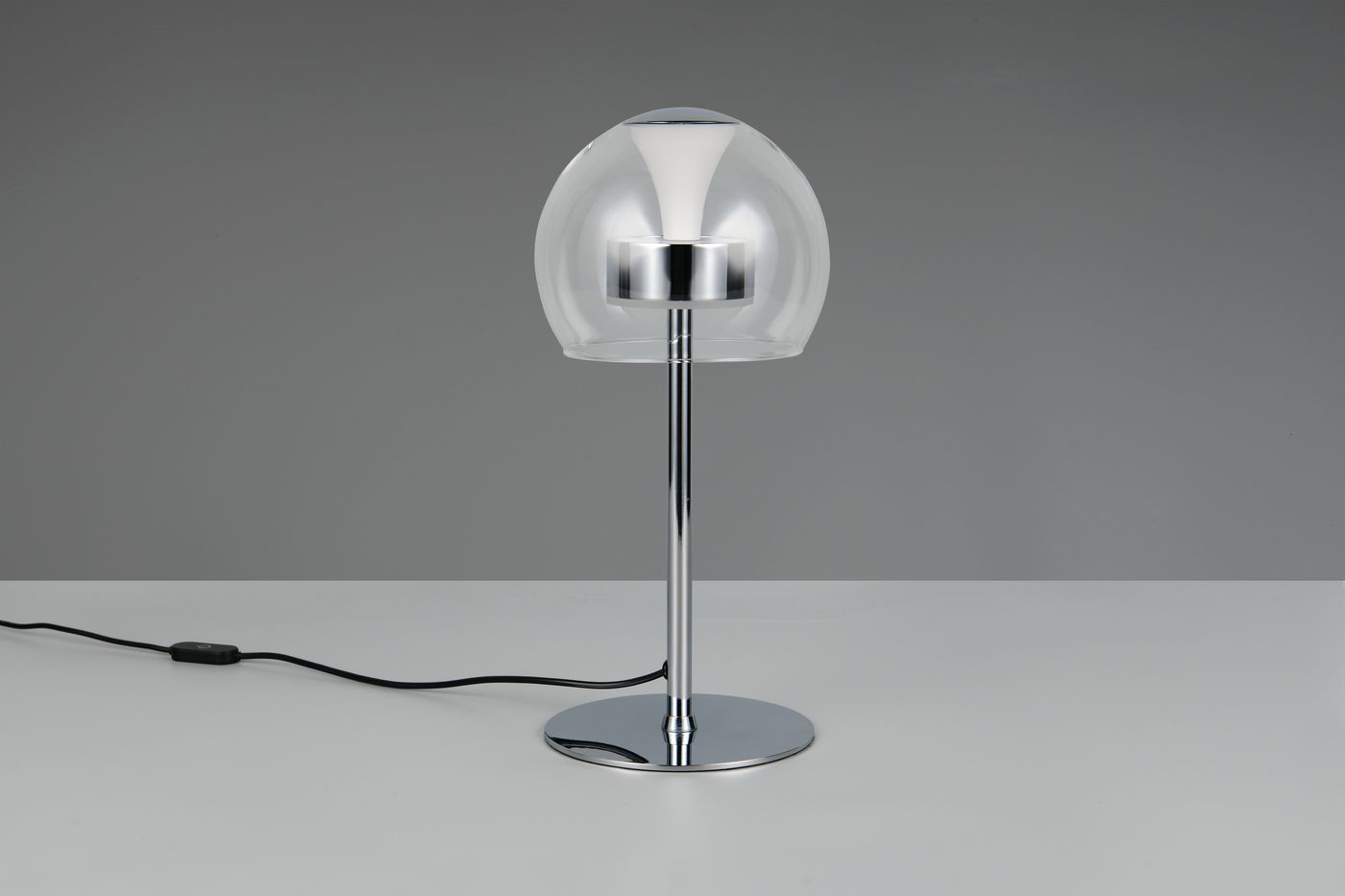 Lampa stołowa Flynt z chromowanym stelażem i przezroczystą kloszem z tworzywa. Ujęcie frontalne, produkt stojący na białym blacie. Widoczny przewód z włącznikiem. Idealna do nowoczesnych wnętrz.