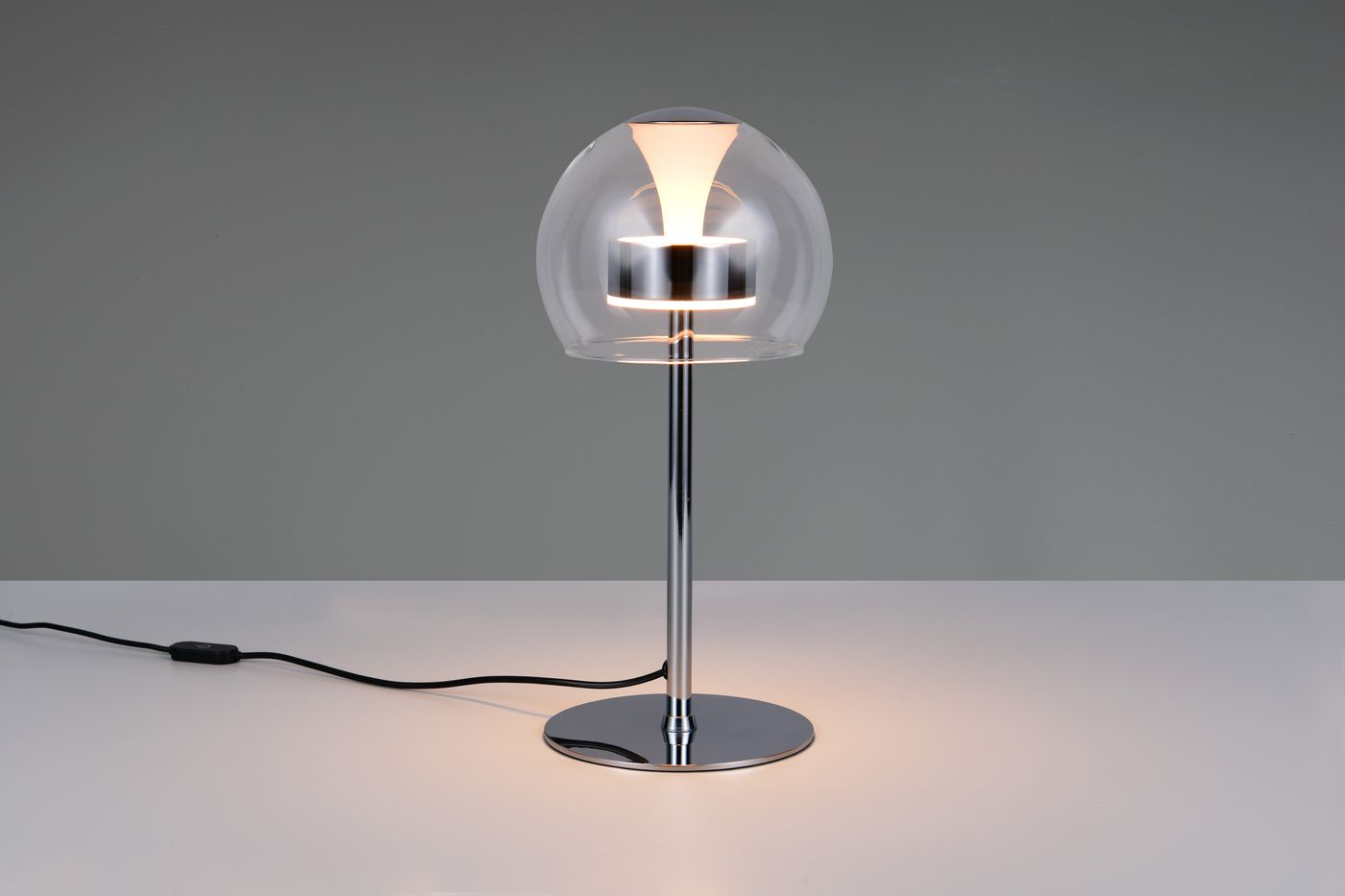 Lampa stołowa Flynt z chromowanym stelażem, szklanym kloszem w kształcie kuli oraz źródłem światła LED 8W. Ujęcie frontalne, produkt stoi na białym blacie, widoczny kabel z włącznikiem. Idealna do nowoczesnych wnętrz.