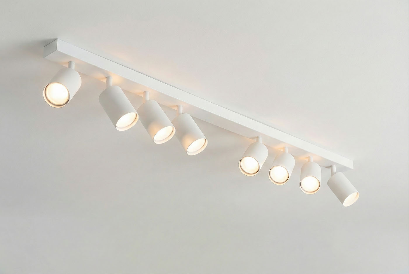 Biała lampa sufitowa typu spot z ośmioma punktami świetlnymi, zamontowana na prostokątnej belce. Reflektory mają cylindryczny kształt i są skierowane w różnych kierunkach, co umożliwia regulację oświetlenia w pomieszczeniu.