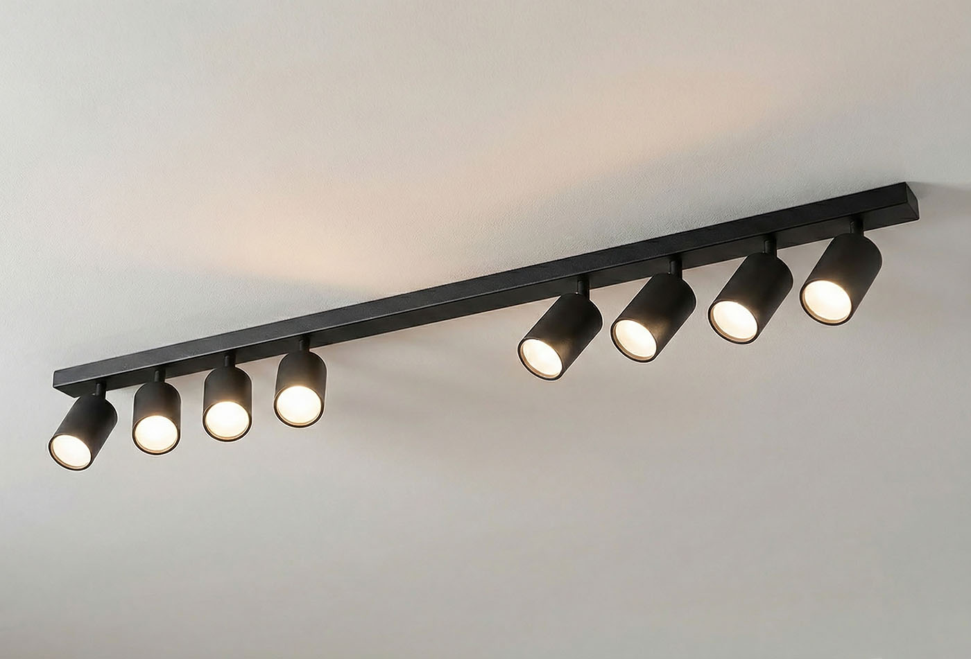 Czarna lampa sufitowa typu spot z ośmioma punktami świetlnymi, zamontowana na prostokątnej belce. Ujęcie z góry, ukazujące równomierne rozmieszczenie reflektorów o cylindrycznym kształcie. Idealna do oświetlenia wnętrz.