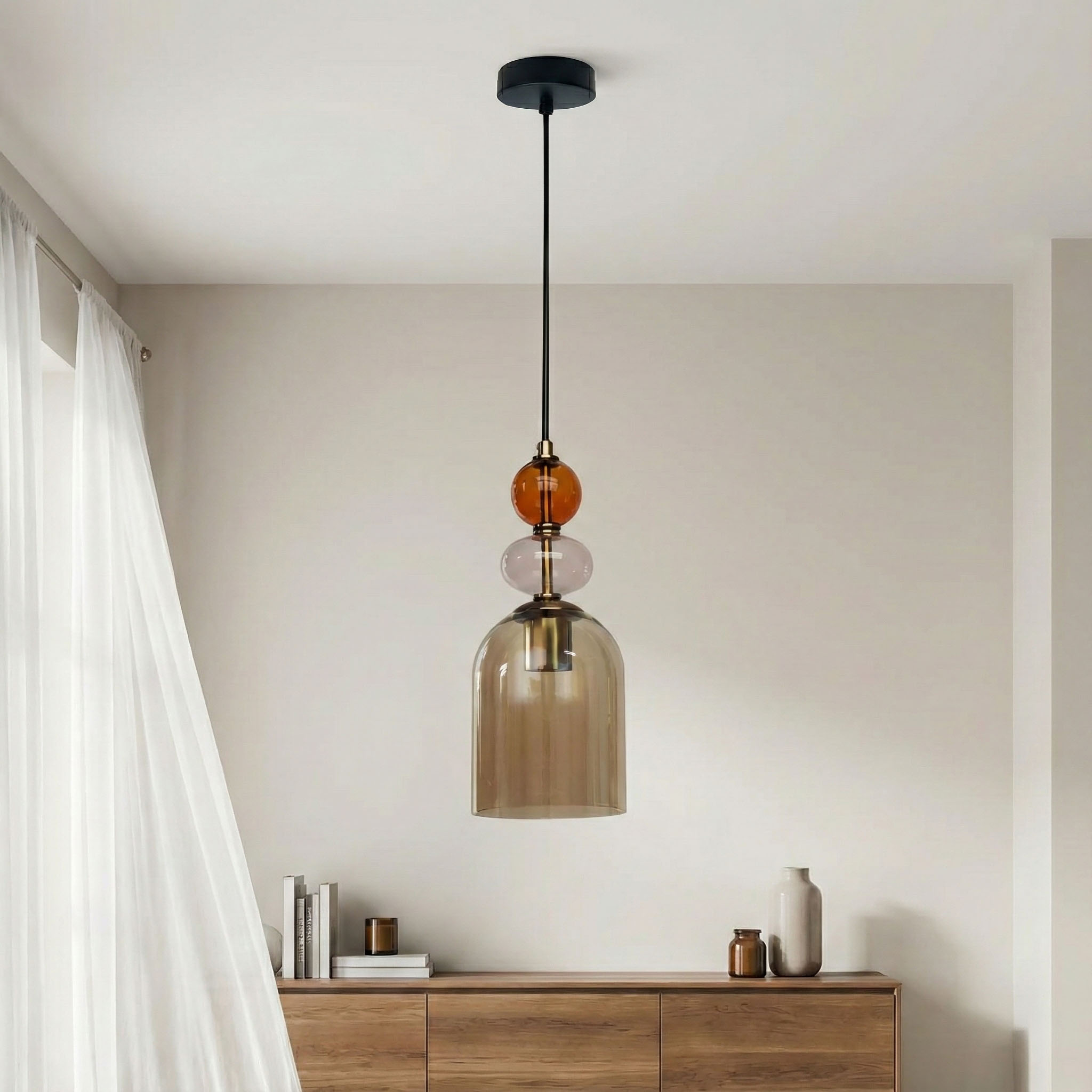 Lampa wisząca Tropea z jedną oprawą E27, z czarnym stelażem i bursztynowymi oraz przezroczystymi szklanymi detalami. Zawieszona na cienkim kablu, z kloszem o kształcie dzwonu, w minimalistycznym wnętrzu z drewnianą komodą.