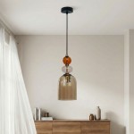 Lampa wisząca Tropea z jedną oprawą E27, z czarnym stelażem i bursztynowymi oraz przezroczystymi szklanymi detalami. Zawieszona na cienkim kablu, z kloszem o kształcie dzwonu, w minimalistycznym wnętrzu z drewnianą komodą.