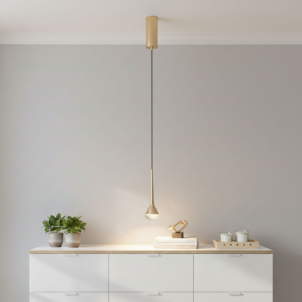 Lampa wisząca Crima Gold LED 1-punktowa w złotym kolorze z czarnym przewodem, zawieszona na białym tle. Kształt w formie kropli z przezroczystym kloszem na końcu. Produkt umieszczony nad białą komodą z roślinami doniczkowymi obok.