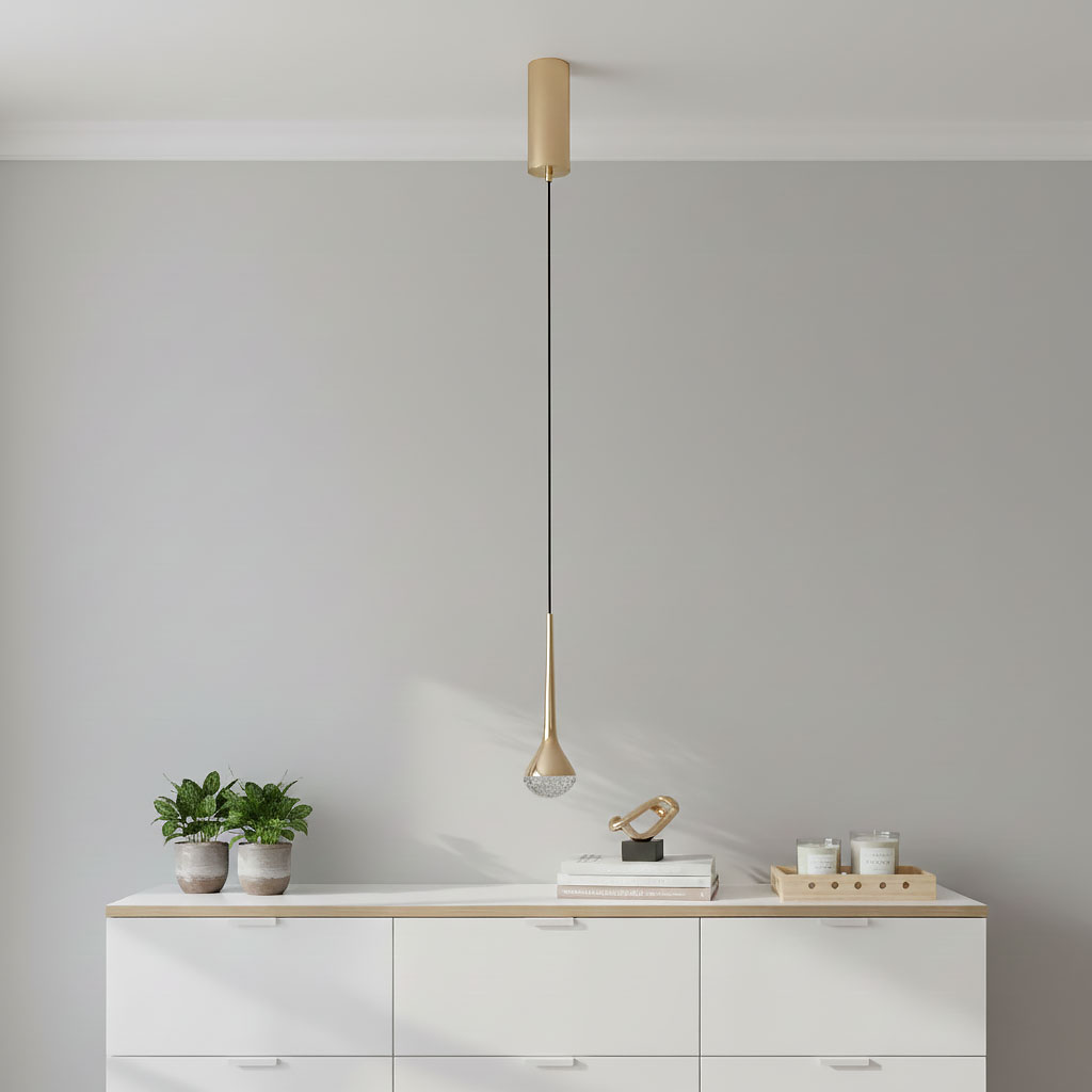 Złota lampa wisząca Crima Gold LED o kształcie kropli, zawieszona na czarnym przewodzie. Elementy metalowe w kolorze złotym, przezroczysta część u dołu lampy z diodami LED. Produkt w nowoczesnym stylu, umieszczony nad białym meblem we wnętrzu.