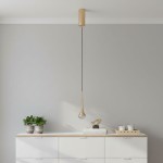 Złota lampa wisząca Crima Gold LED o kształcie kropli, zawieszona na czarnym przewodzie. Elementy metalowe w kolorze złotym, przezroczysta część u dołu lampy z diodami LED. Produkt w nowoczesnym stylu, umieszczony nad białym meblem we wnętrzu.