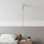 Lampa wisząca Crima Cromo LED w kształcie kropli, chromowane wykończenie, zawieszona na cienkim przewodzie. Umieszczona nad stolikiem nocnym w sypialni. Tło w odcieniach szarości i bieli, z widoczną rośliną doniczkową obok.
