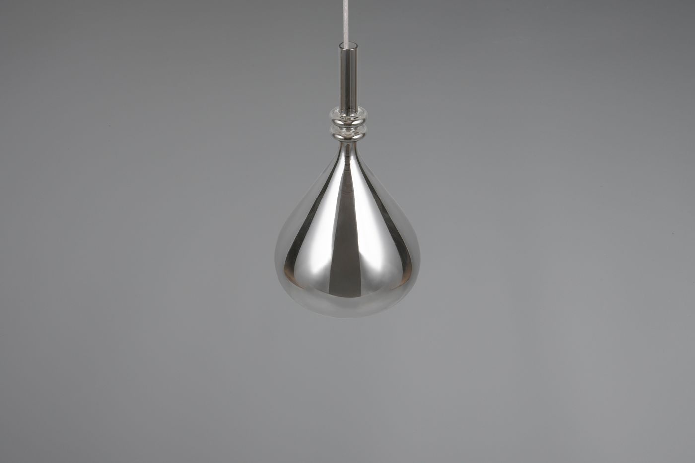 Lampa wisząca Aldea w kolorze chromu, o kształcie kropli. Ujęcie frontalne, zwisająca z cienkiego, białego przewodu. Gładka, błyszcząca powierzchnia. Przeznaczona na żarówkę E14. Tło szare, jednolite.