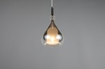 Lampa wisząca Aldea z kloszem w kształcie kropli, wykonana z przezroczystego szkła i chromowanej stali. Zawieszona na cienkim, białym przewodzie. Źródło światła E14, emituje ciepłe światło, umieszczone w środkowej części klosza. Tło w odcieniu szarości.