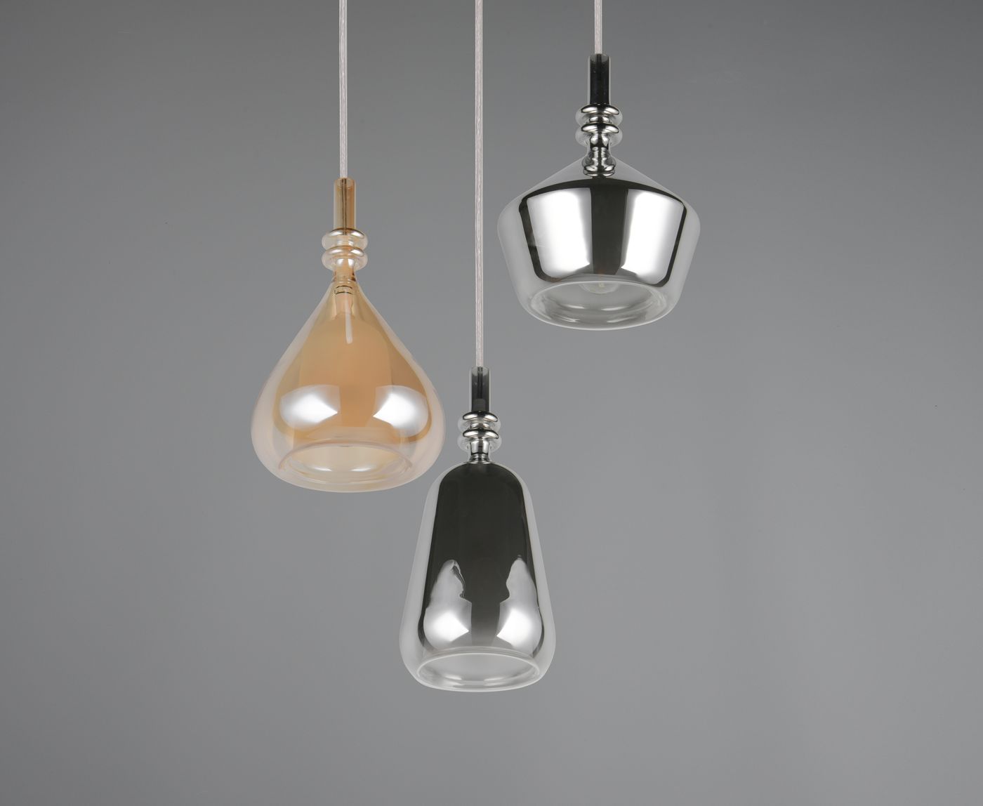 Lampa wisząca Aldea 3xE14 w kolorze białym i multikolorowym, składająca się z trzech kloszy w różnych kształtach: kroplowaty w odcieniu pomarańczowym, cylindryczny metaliczny oraz szklany w kolorze czarnym. Zawieszona na białych linkach, na szarym tle.
