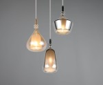 Lampa wisząca Aldea z trzema kloszami E14 w różnych kształtach: przezroczysty, złoty i metaliczny. Zawieszona na cienkim przewodzie, idealna do nowoczesnych wnętrz. Klosze emitują ciepłe światło, tworząc przytulną atmosferę. Tło szare.