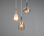 Lampa wisząca Aldea z trzema kloszami w różnych kształtach, w kolorach białym i metalicznym. Klosze zawieszone na białych przewodach, emitują ciepłe światło. Tło w odcieniu szarym, podkreślające elegancję lampy.