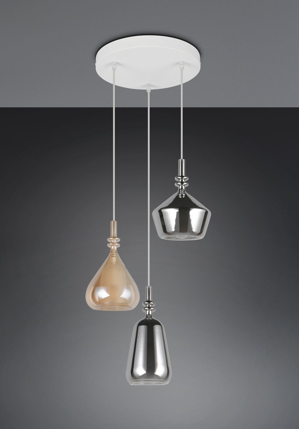 Lampa wisząca Aldea z trzema kloszami w kształcie kropli, w kolorze przezroczystym i metalicznym. Klosze zawieszone na białych linkach, montowane do okrągłej, białej podsufitki. Ujęcie frontalne, z jasno szarym tłem.