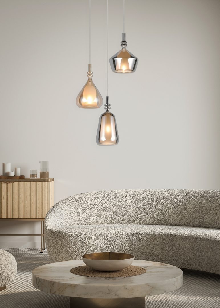 Lampa wisząca Aldea z trzema źródłami E14, w białym kolorze i multikolorowych kloszach. Ujęcie z przodu, z lampami w różnych kształtach: kropli, stożka i cylindrycznym. Produkt zawieszony nad okrągłym stołem na tle jasnej ściany.