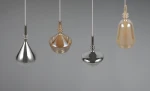 Lampa wisząca Aldea z czterema kloszami w różnych kształtach: metaliczny srebrny, przezroczysty, złoty i matowy. Powieszona na białych przewodach, z widocznymi oprawkami E14. Idealna do nowoczesnych wnętrz. Tło szare, minimalistyczne.