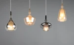 Lampa wisząca Aldea z 4 kloszami E14 w różnych kształtach i kolorach, w tym przezroczystym, złotym i metalicznym. Zawieszona na białych kablach, w aranżacji na szarym tle, prezentuje nowoczesny design.