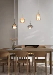 Lampa wisząca Aldea z czterema kloszami E14 w białym i multikolorowym wykończeniu. Klosze o różnych kształtach zawieszone na długich linkach, umieszczone nad drewnianym stołem w nowoczesnym wnętrzu. Tło jasne, minimalistyczne.
