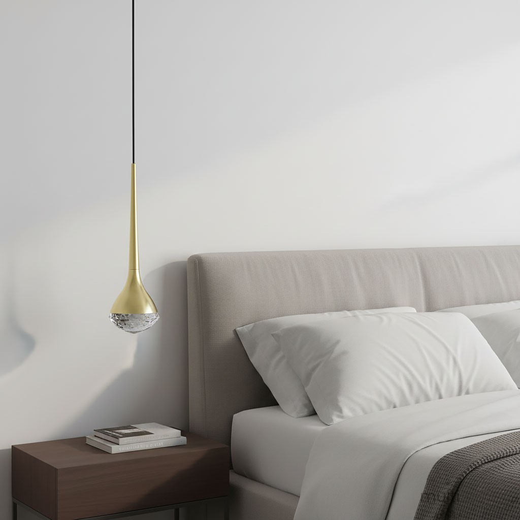 Lampa wisząca LED Crima Old Gold 6W 3000K w kolorze złota satynowego, w kształcie kropli, zawieszona nad łóżkiem. Posiada przezroczysty klosz na dolnej części, zamontowana na cienkim czarnym przewodzie. Tło jasne, z elementami sypialni.