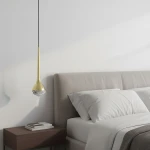 Lampa wisząca LED Crima Old Gold 6W 3000K w kolorze złota satynowego, w kształcie kropli, zawieszona nad łóżkiem. Posiada przezroczysty klosz na dolnej części, zamontowana na cienkim czarnym przewodzie. Tło jasne, z elementami sypialni.