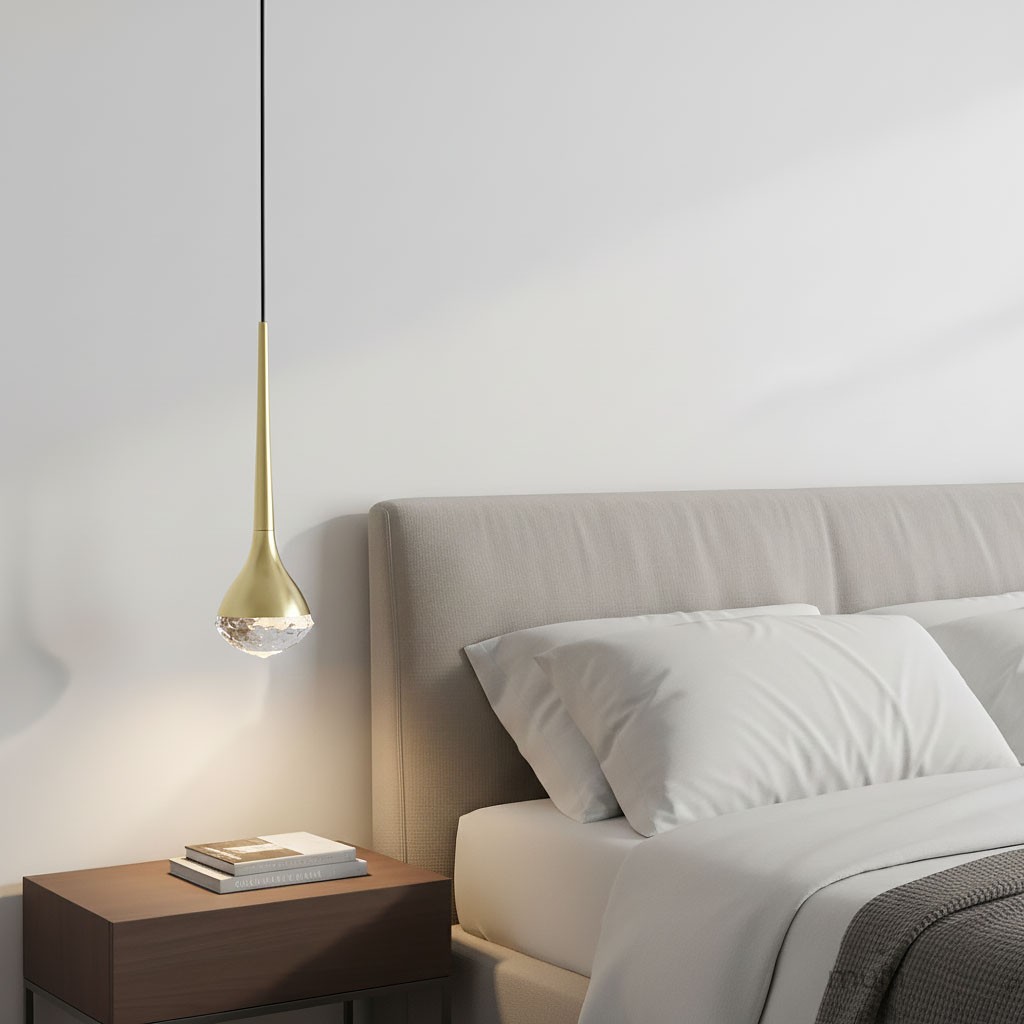 Lampa wisząca LED Crima Old Gold o mocy 6W, barwie światła 3000K. Złoto satynowe, smukły kształt z przezroczystym detalem u dołu. Zawieszona nad łóżkiem, w nowoczesnym wnętrzu, na tle jasnej ściany i szarej pościeli.
