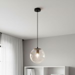 Lampa wisząca Sarpo z czarnym uchwytem, zawieszona na cienkim przewodzie. Szklany klosz w formie kuli, przezroczysty, z widoczną żarówką E27 w środku. Tło w neutralnym kolorze, minimalistyczne wnętrze z białymi meblami.