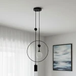 Lampa wisząca Trosti 2-punktowa, czarna. Konstrukcja z okrągłym elementem oraz szklanym kloszem w kształcie kuli. Dwa przewody zawieszone w pionie, zamontowana na suficie. Tło w jasnym kolorze z zasłonami i obrazem w ramie.